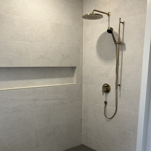 A modern shower with a rainfall showerhead and a handheld shower-head on a brushed brass fixture against a light gray tiled shower wall with a built-in niche.